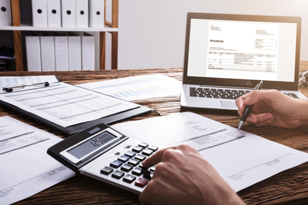 Close-up Of A Businessperson's Hand Calculating Bill With Laptop On Wooden Desk