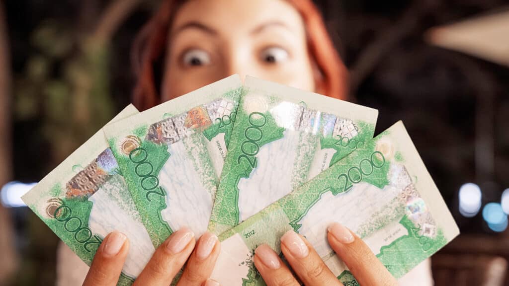 Surprised young woman holding a fan of Kazakhstan tenge banknotes in front of her face, wide-eyed expression