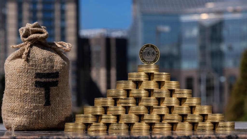 Pyramid of coins in denominations of 100 Kazakhstani tenge and a money bag with a symbol of tenge on the background of modern buildings