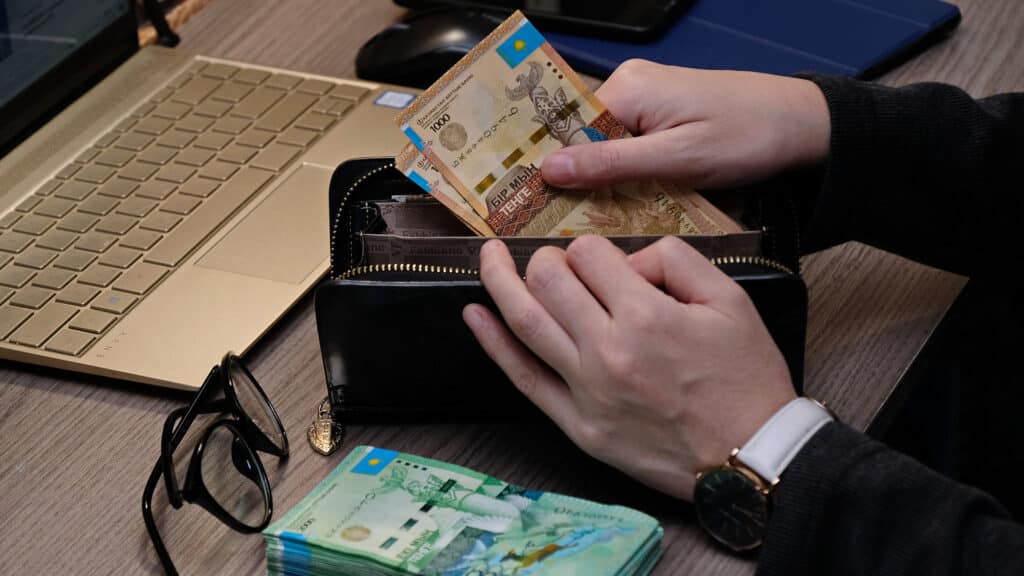 Almaty, Kazakhstan - 12.01.2020 : A person puts Kazakhstani tenge in a wallet while sitting at a table with a computer.