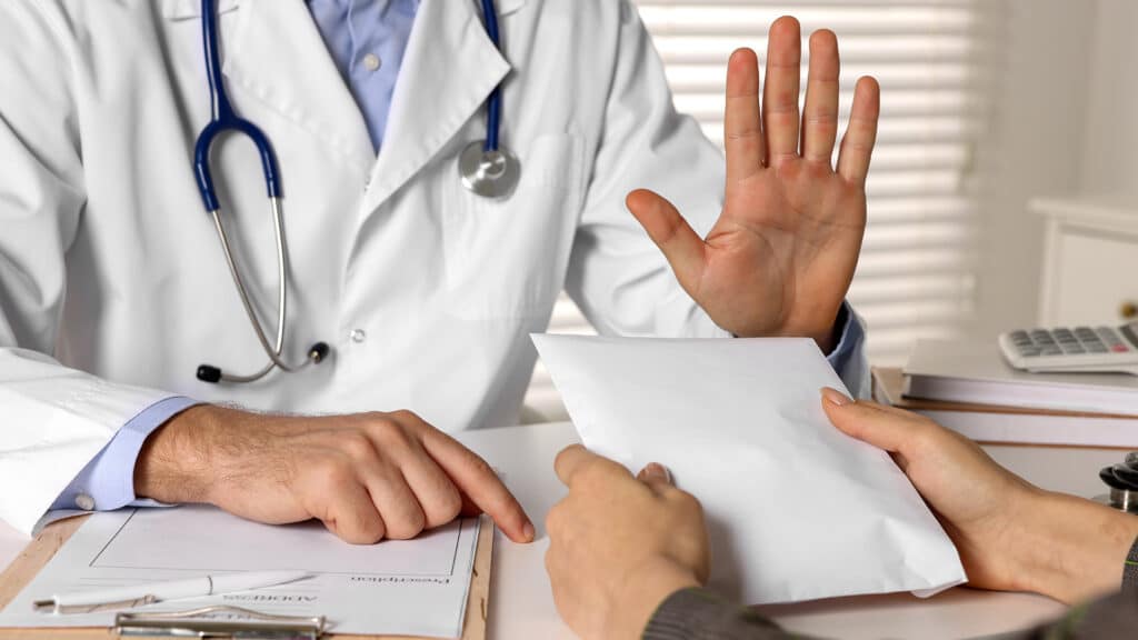 Corruption concept. Woman giving envelope with money to doctor at white table, closeup