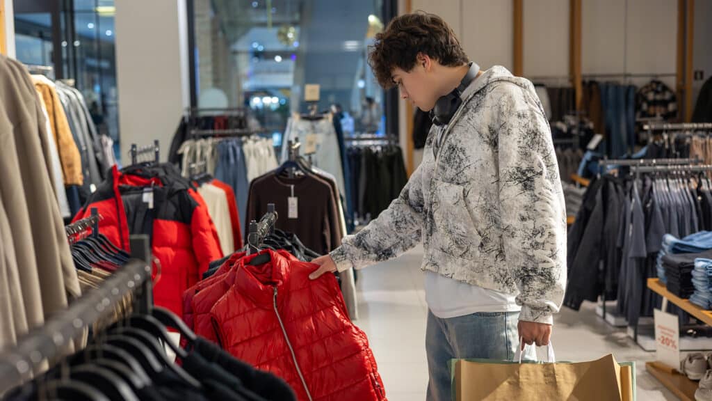 Teenager shopping for winter clothes in fashion store, young man choosing red jacket among casual clothing racks, youth lifestyle and retail concept. youth fashion, shopping habits.