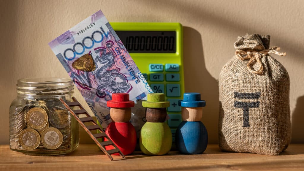 A group of symbolic wooden people near a glass jar with motets in denominations of 100 and 200 Kazakhstani tenge and a 10,000 tenge banknote