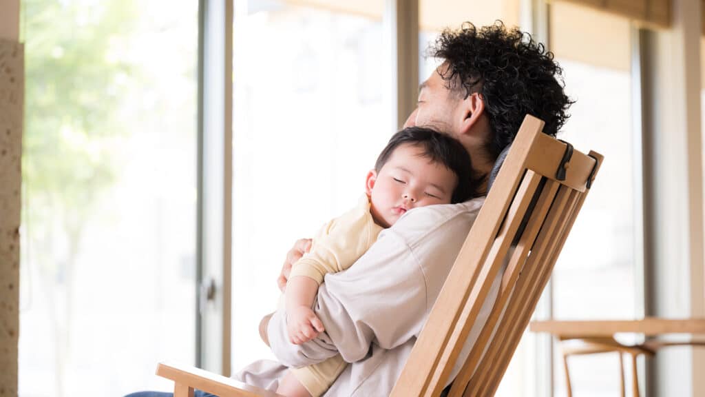 A middle-aged Japanese man sitting in a chair holding a baby. The image of a father putting his child to sleep.