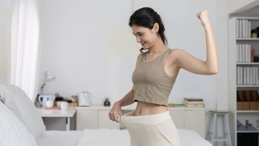 Happy women flexing arm while wearing oversized pants, celebrating successful weight loss. Concept of fitness, diet achievement, body transformation, and healthy lifestyle.