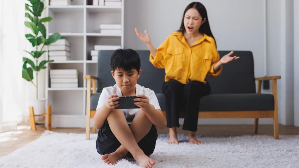 Asian mother scolding her son for playing video games on a mobile phone while sitting on the living room floor, creating a tense moment