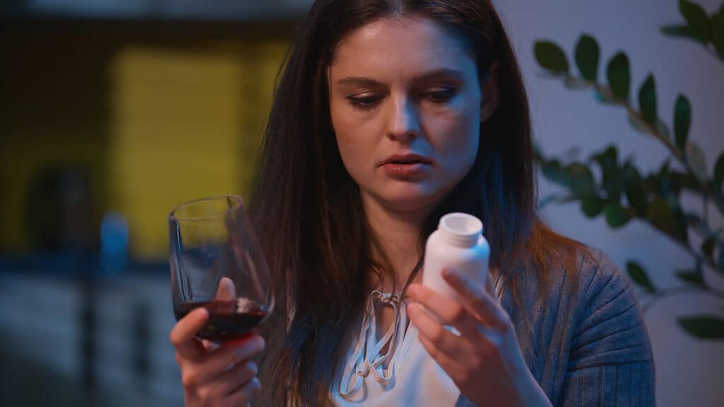 Sad upset woman holding glass wine pills thinking difficult choice sick unwell drink alcohol addiction worried girl drinking antibiotic health problem disease frustrated Caucasian female at home night