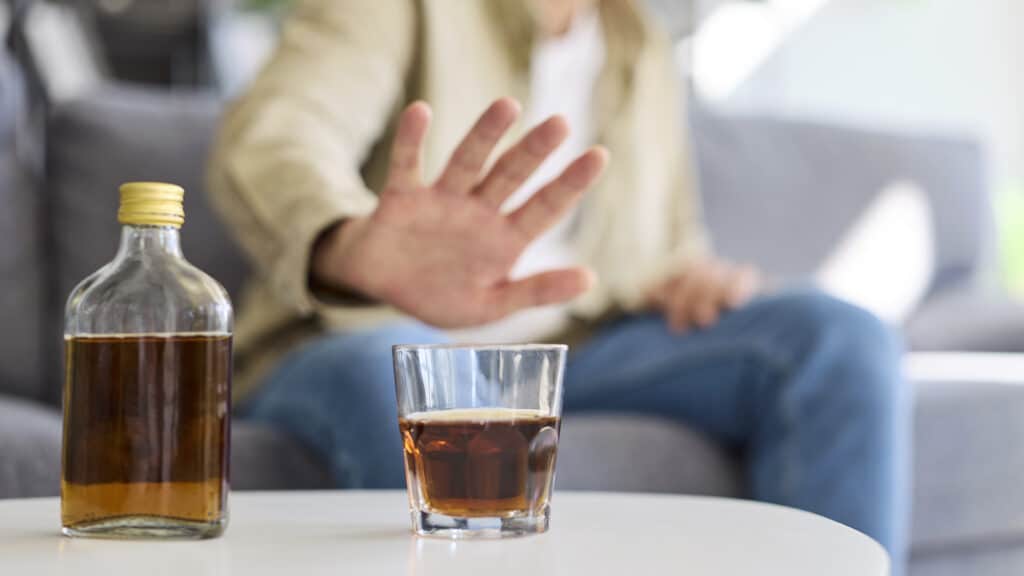Man shows refusal to alcohol, sobriety choice. A man raises a stop hand to a glass at home, showing abstinence resisting temptation choosing health amid addiction. Theme recovery over drinking.