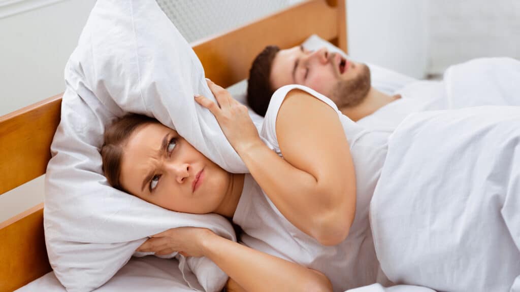 Snoring. Angry Girlfriend Covering Ears With Pillow Lying Near Sleeping Boyfriend In Bed At Home. Selective Focus