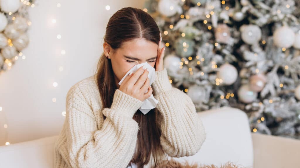A young woman sneezing while sitting on a sofa, wrapped in a warm blanket in front of a decorated Christmas tree.