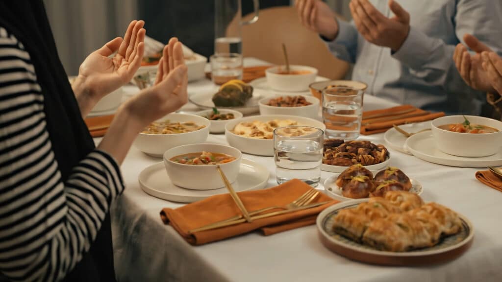 A Muslim family shares a traditional Iftar meal, with hands lifted in gratitude and prayer before eating
