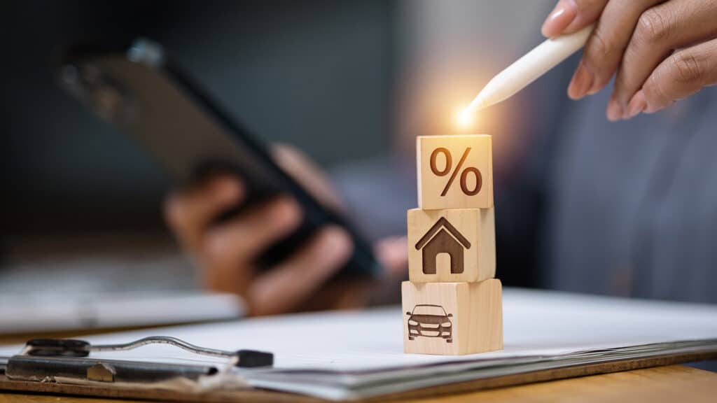 Man using smartphone and stylus next to loan documents while wooden blocks show car, house and percentage. Concept of interest rate, home loan and car loan approval, refinancing, budgeting.