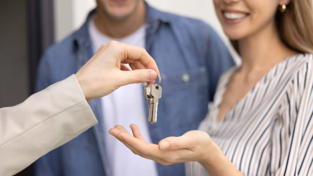 Close up cropped view of female realtor handing over house keys to smiling young couple, purchasing, renting, or leasing property. Homeownership, real estate transactions, and financial investments