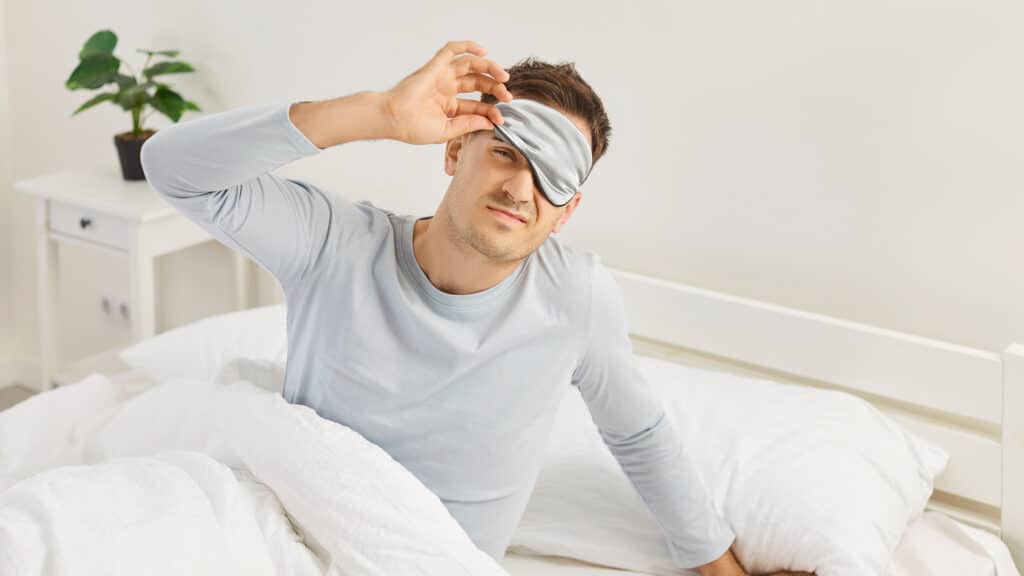 Man who hates weekday morning routine and has not had enough sleep sits on bed, raises eye mask, looks at camera with displeased sleepy bad mood face expression with no energy to wake up to start week
