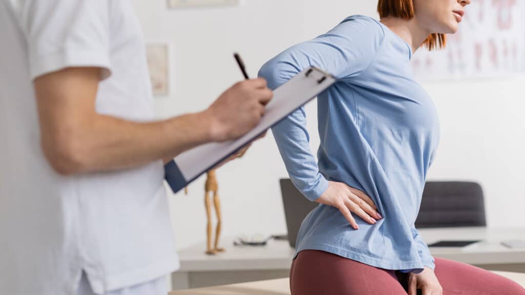 cropped view of physiotherapist with clipboard writing diagnosis near woman suffering from pain in loin in consulting room