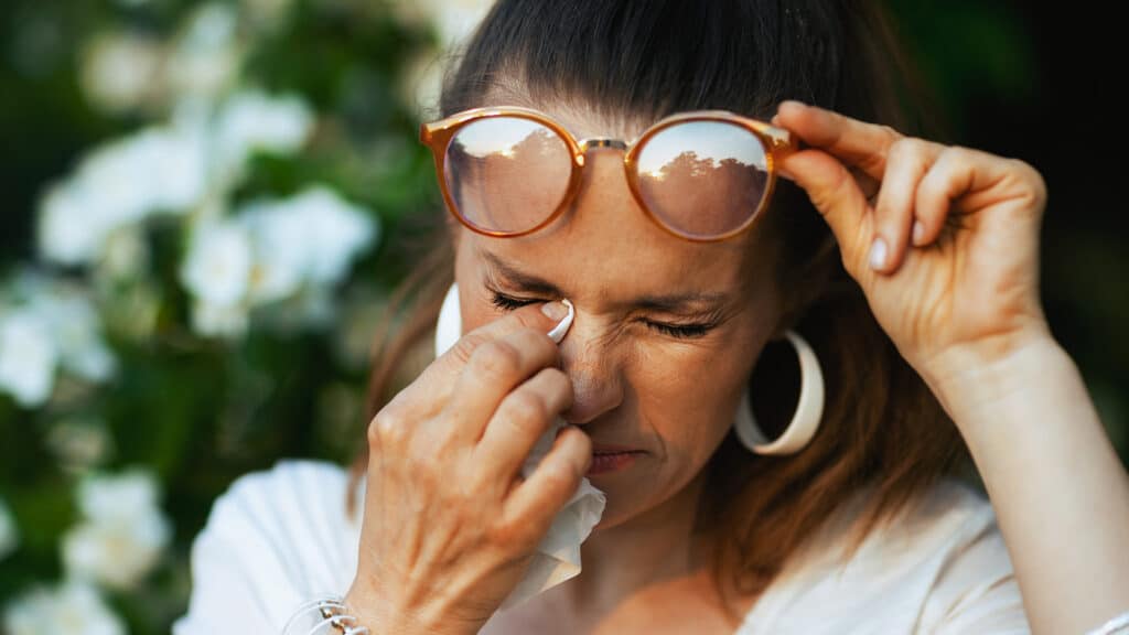 Summer time. sad modern 40 years old woman in white shirt with handkerchief and eyeglasses has an allergy attack near flowering tree.