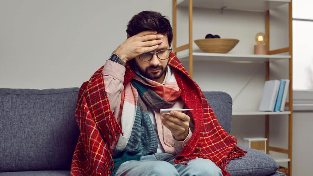 Sick man with fever looks confusedly at thermometer showing high body temperature. Worried young man wrapped in plaid grabs his head while sitting on sofa at home. Concept of viral infection.