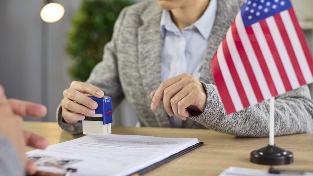 US visa documents approval by consulate officer at office desk with United States flag. Lawyer holding stamp to approve USA immigration application, paperwork and bureaucracy process at interview
