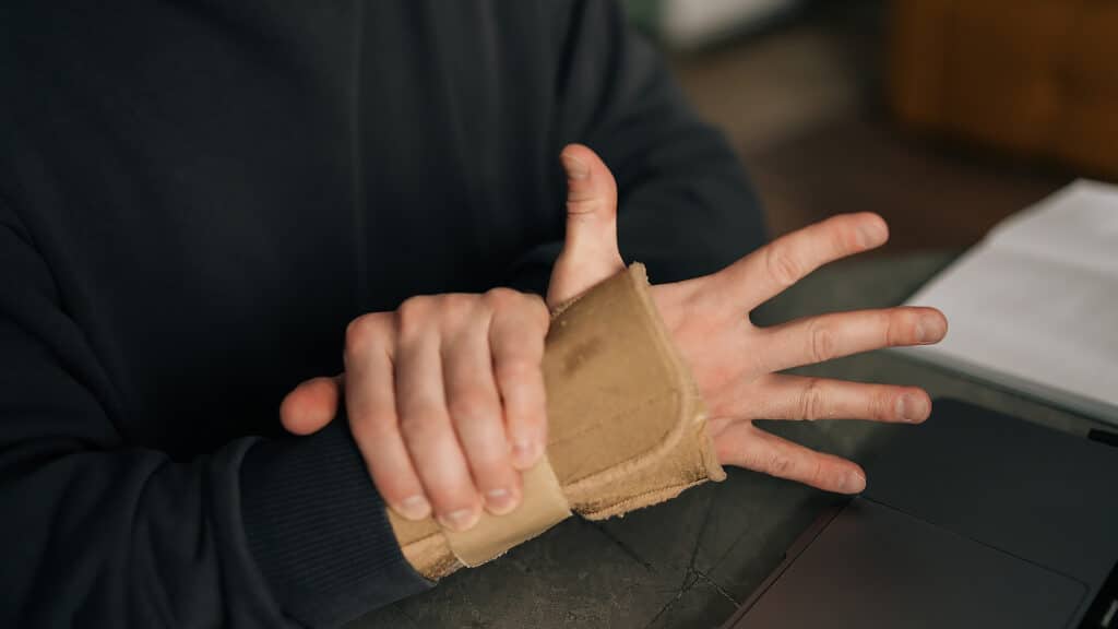 Close-up cropped shot of male hands wearing wrist brace, massaging painful wrist while working remotely from home office, surrounded by laptop and notebook. Concept of struggles of remote work