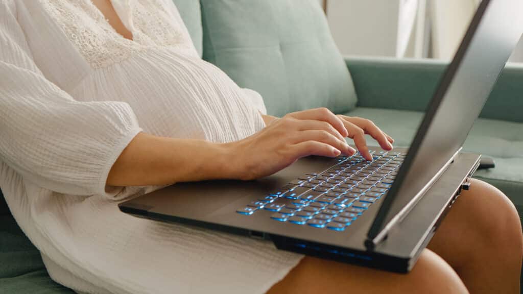 Midsection shot of unrecognizable pregnant woman working on laptop placed on laps doing paperwork at home.. Pregnant woman working at computer. Freelance and remote work.