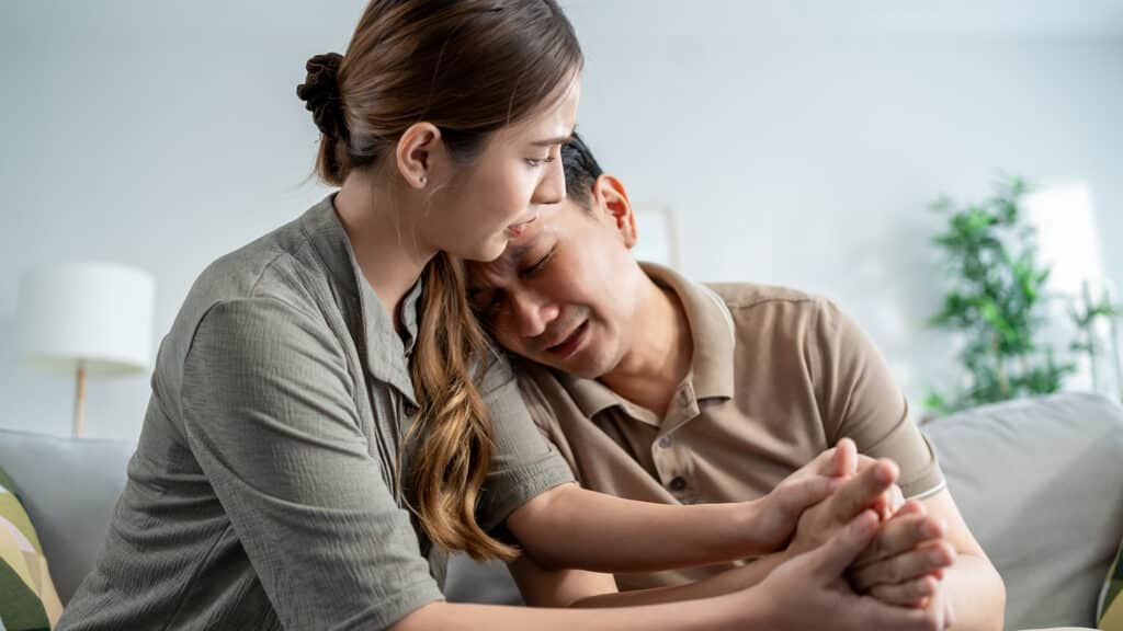 Asian wife comforting upset husband crying with tears for house problem. Attractive woman support consoling and understand empathy to depressed stressed man in living room at home. Family relationship
