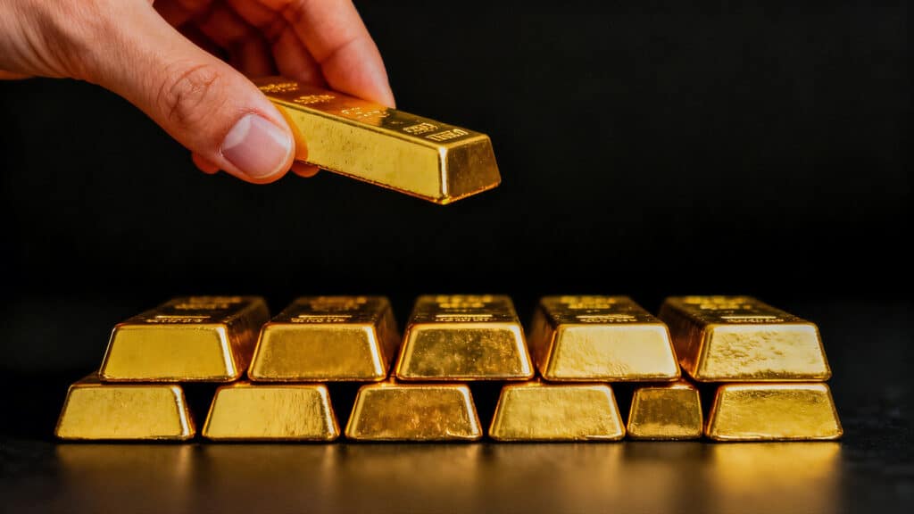 Hand holding gold bar over stack of gold bars on black reflective surface with dark background