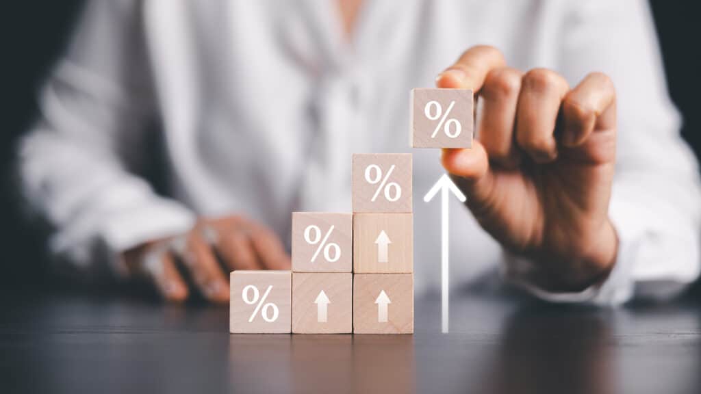 Business hand placing percent block on rising wooden staircase with upward arrows, symbolizing interest rate hikes, inflation pressure, higher loan costs and planning decisions. interest rate hikes