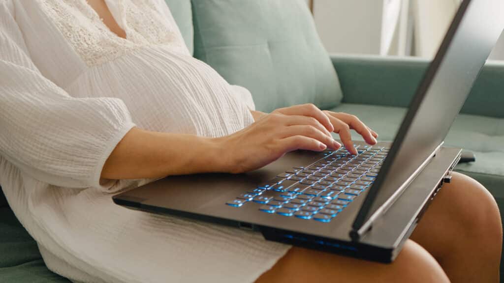 Midsection shot of unrecognizable pregnant woman working on laptop placed on laps doing paperwork at home.. Pregnant woman working at computer. Freelance and remote work.