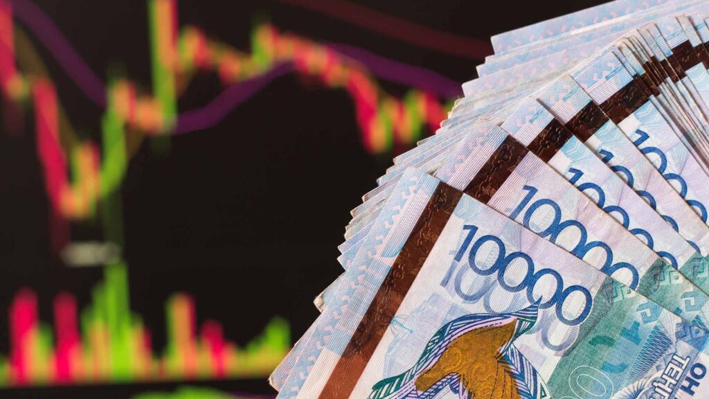Tenge in Kazakhstan against the background of a laptop with an open chart of the currency market or stock exchange. Hands holding Tenge in front of the computer. Tenge KZT, bank of Kazakhstan.