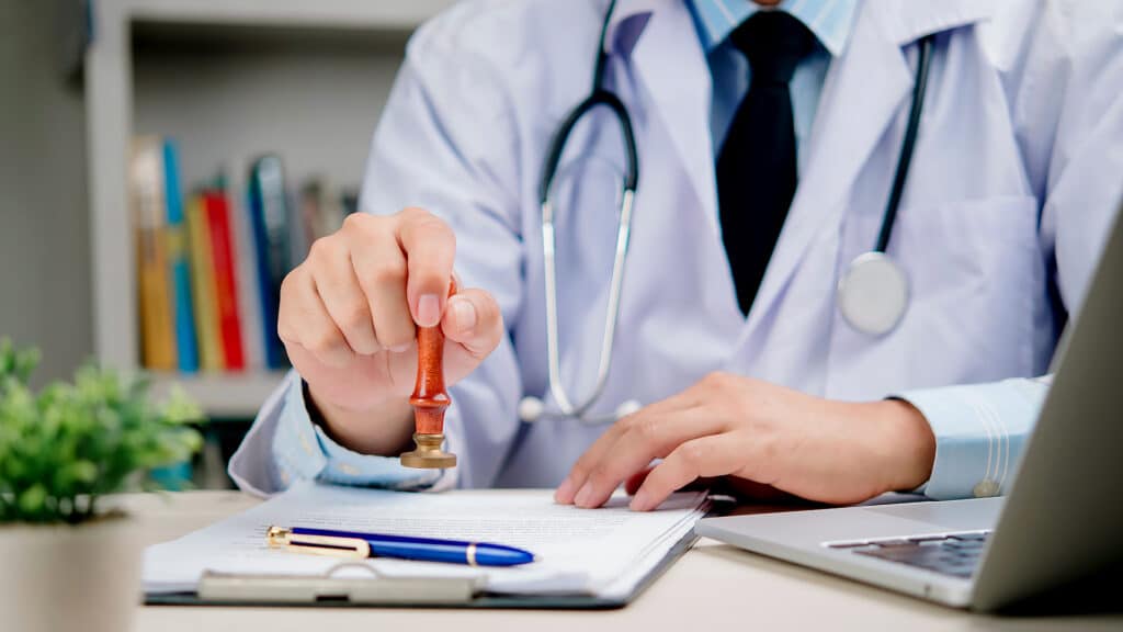 Doctor white coat with stethoscope stamping medical document for approval. Concept for health certificate, insurance claim, diagnosis validation, and healthcare paperwork hospital or clinic office.
