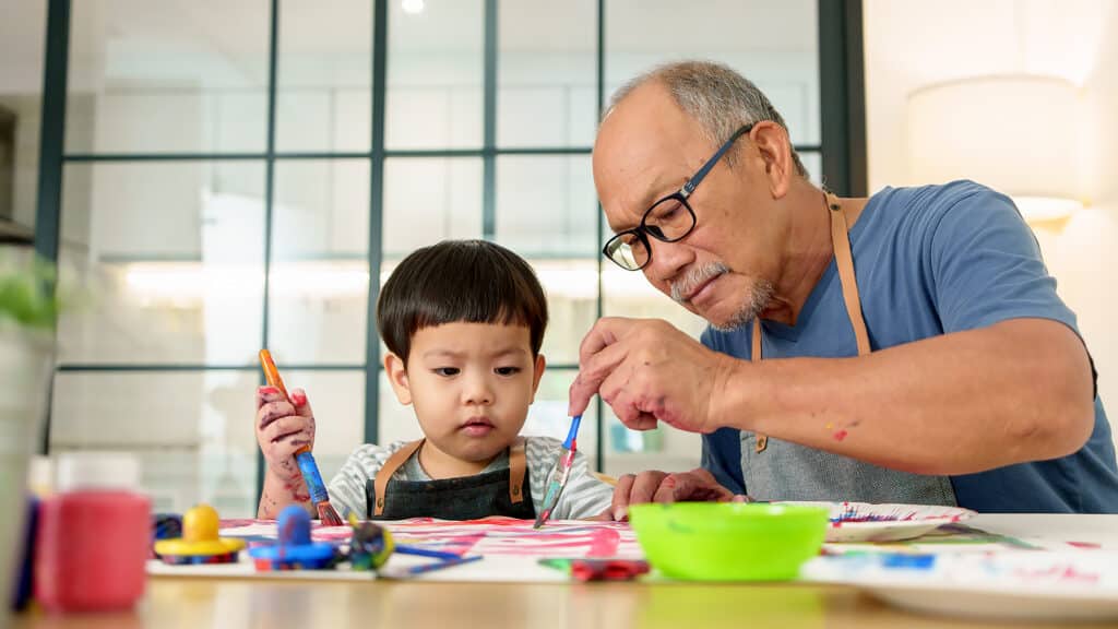 Asian Senior man and young child or grandchild painting together at a table, displaying creativity and focus in a nurturing family environment.