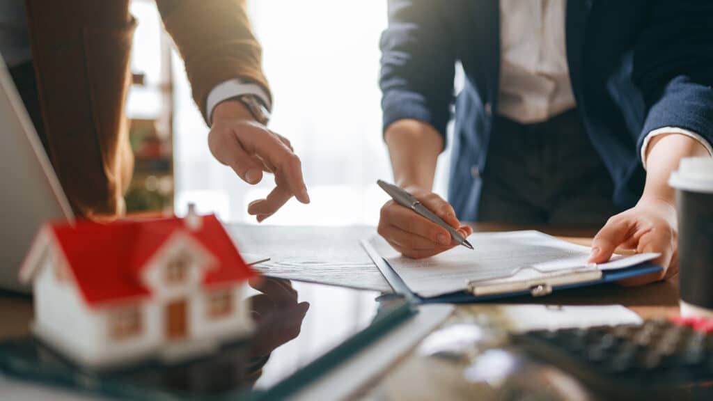 Professional business partners negotiating and signing a real estate contract. Bright, hopeful atmosphere with hands pointing and writing on documents, symbolizing trust and collaboration. A model house on the desk represents homeownership dreams, investment, and the personal journey of buying a home.