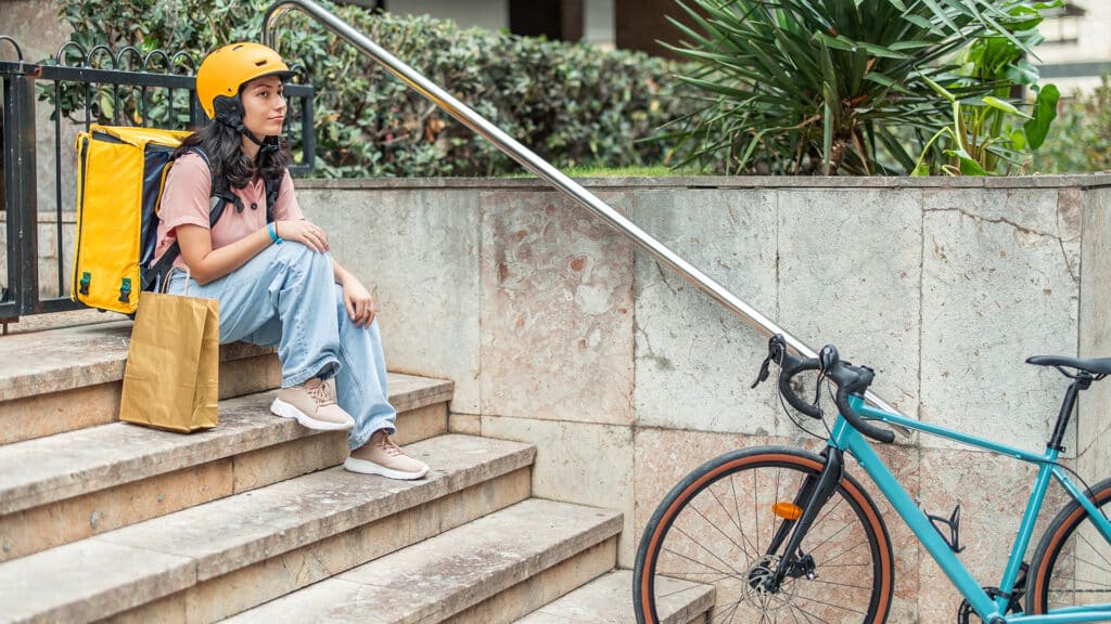 Young Asian woman courier wearing a yellow helmet and backpack sitting on urban steps next to her bicycle, holding a paper bag and waiting for the next food delivery