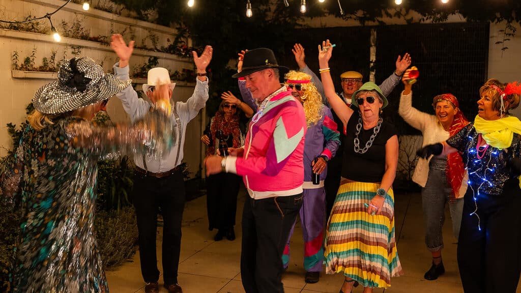 A diverse group of elderly individuals is seen having a joyous time at an outdoor costume party. The attendees, both men and women, are dressed in colorful and extravagant costumes, including cowboy