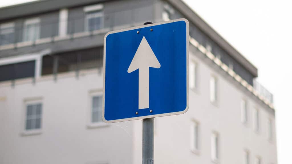 One-way street sign in Germany. The white arrow on the blue square is pointing ahead. The travel direction is straight on.