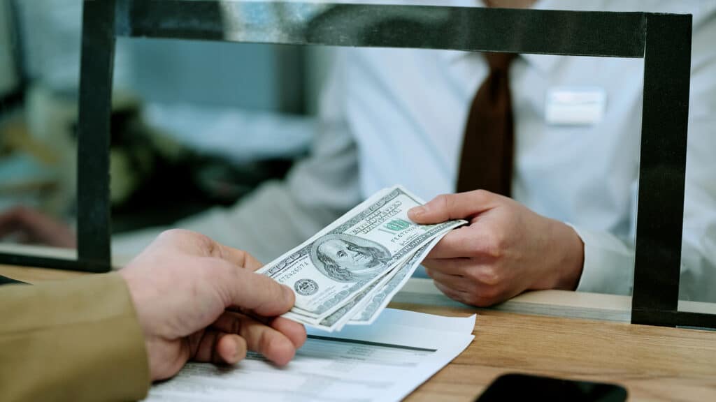 Female bank teller handing cash to Caucasian middle aged man customer through counter window, both interacting during financial transaction at bank branch