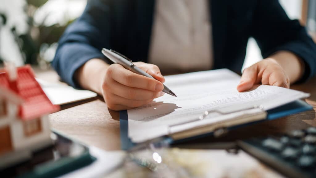 Professional businesswoman signing important real estate documents at a modern office desk, conveying trust, reliability, and attention to detail. The image showcases a close-up of hands with a pen, focused on paperwork, with a miniature house model in the foreground, emphasizing themes of property purchase, legal agreements, and financial planning. The warm, natural lighting and professional setting evoke feelings of confidence, security, and success.