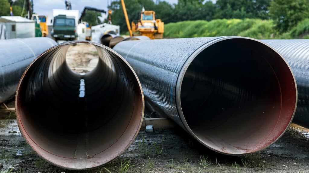 Close-up view of large diameter steel pipes stored on a construction site before installation during a major energy infrastructure project with heavy machinery visible in the background.